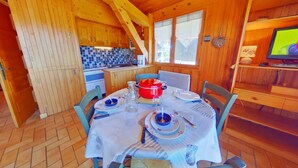 Interior em madeira com uma mesa de jantar posta para quatro, área de cozinha e uma televisão sobre uma prateleira de madeira.