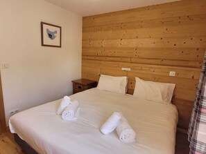 A hotel room with a wooden headboard, a bed with white linens, two rolled towels on the bed, a small wooden nightstand, and a framed picture on the wall. - Modern apartment for 8 with WIFI and parking, Les Gets (Les Gets)