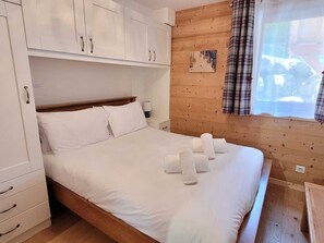 Een slaapkamer met een houten hoofdbord, een bed met witte lakens, een nachtkastje, een lamp en een raam met gordijnen.