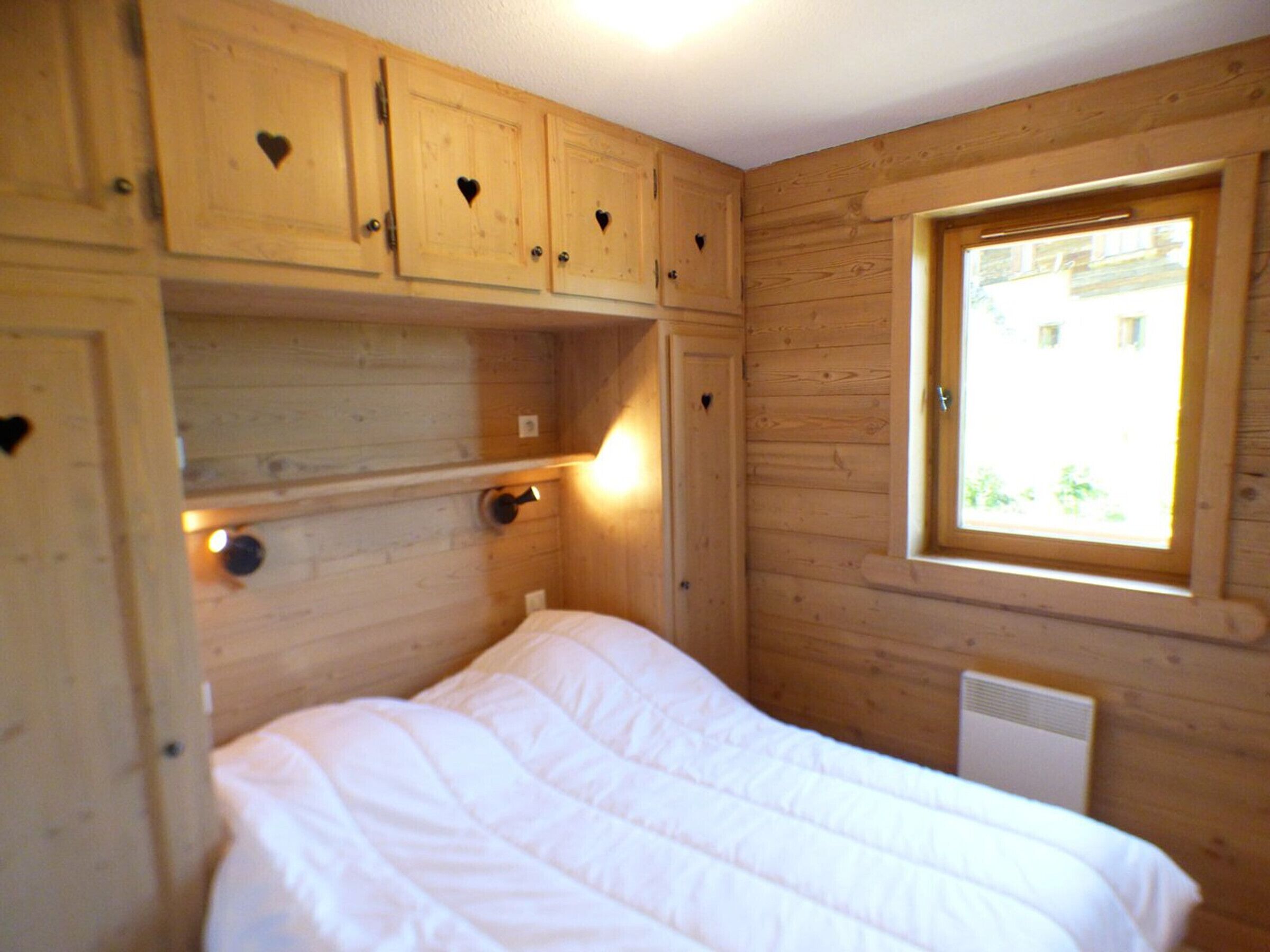 A wooden room with a bed, built-in cabinets with heart-shaped cutouts, a window with a view, and a wall-mounted light.