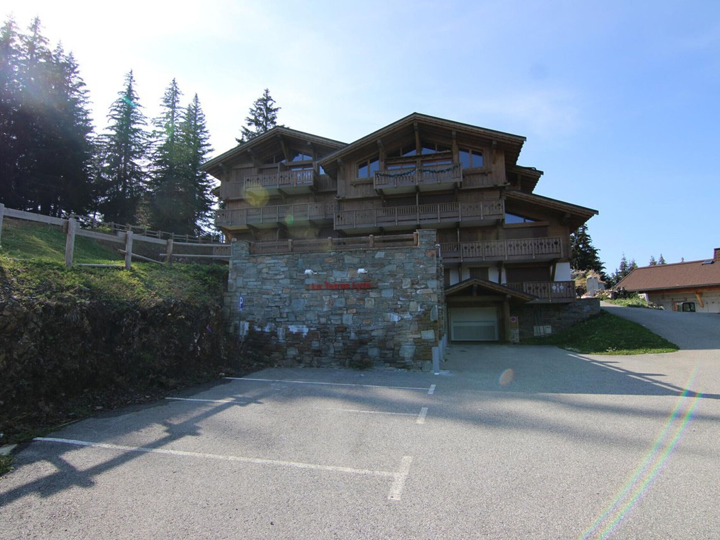 A multi-story wooden building with balconies, a stone wall entrance, and a parking area.