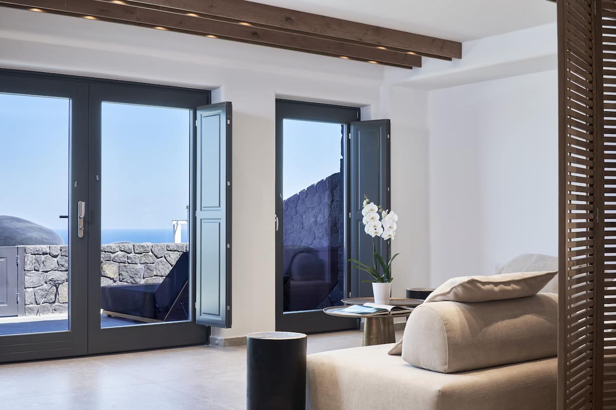 A modern living room with a sofa, a small table, and a potted plant. There are two large sliding doors leading to a balcony with a stone wall and a view of the sea.