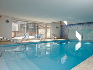 Une piscine intérieure avec des parois en verre transparent, un sol carrelé et trois chaises longues à rayures.