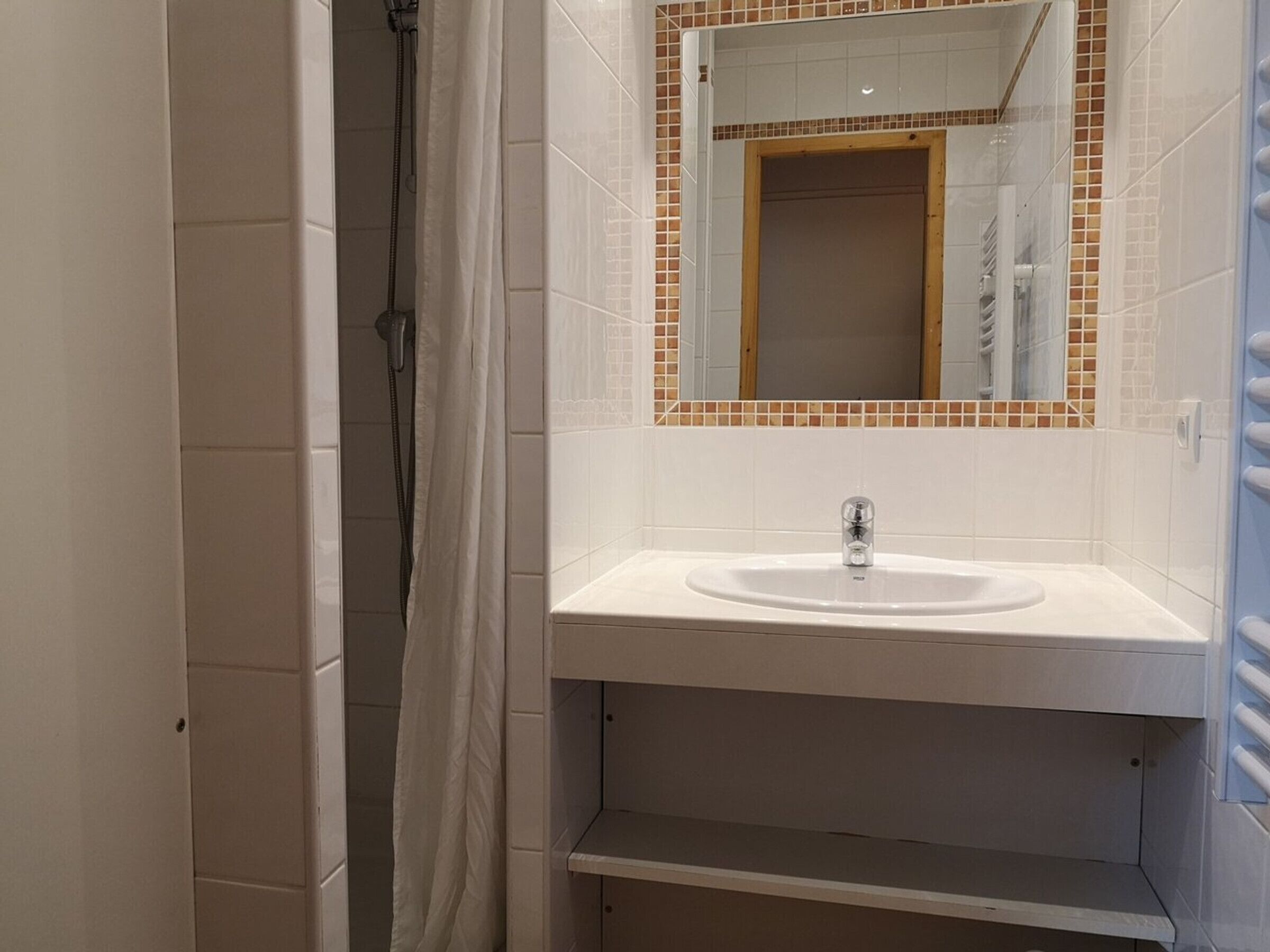 Une salle de bain avec un lavabo blanc, une douche et un miroir dont le cadre est orné de carreaux bruns.
