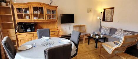 A cozy living room with a dining area, wooden furniture, a flat-screen TV, and a sofa with patterned cushions.