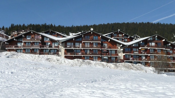 Una hilera de cabañas de madera de varios pisos con balcones, frente a un paisaje nevado y un fondo forestal.