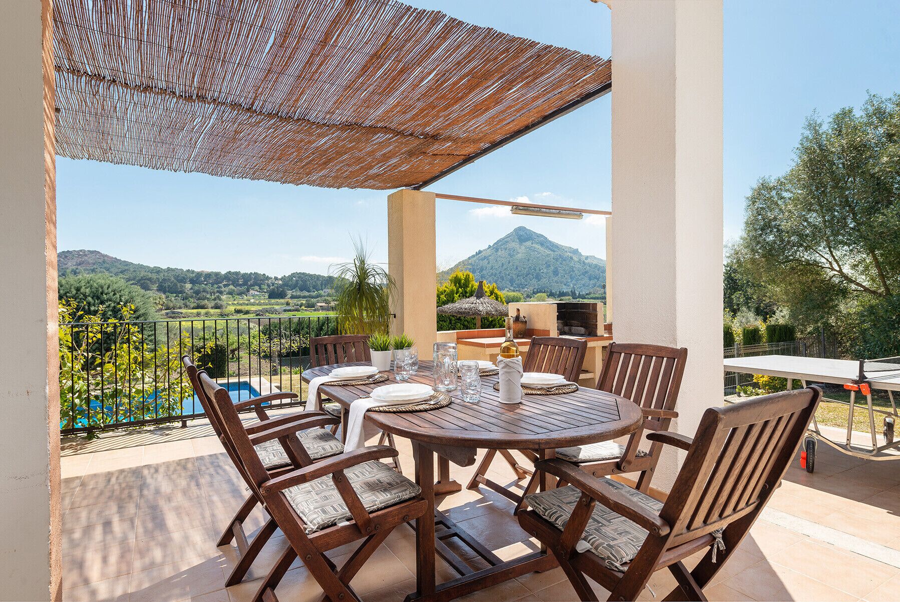 Ein Holztisch mit Stühlen und einem gewebten Sonnenschirm auf einem Balkon mit Blick auf eine Berglandschaft.