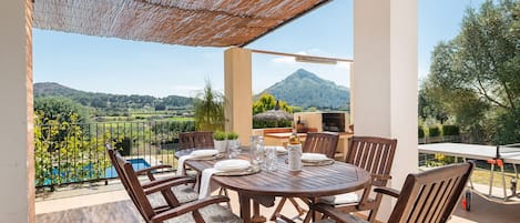 Ein Holztisch mit Stühlen und einem gewebten Sonnenschirm auf einem Balkon mit Blick auf eine Berglandschaft.