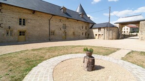 A stone building with a courtyard, a central tree stump with a planter, and a paved walkway. - Large gîte with indoor pool and sauna (Charbogne)