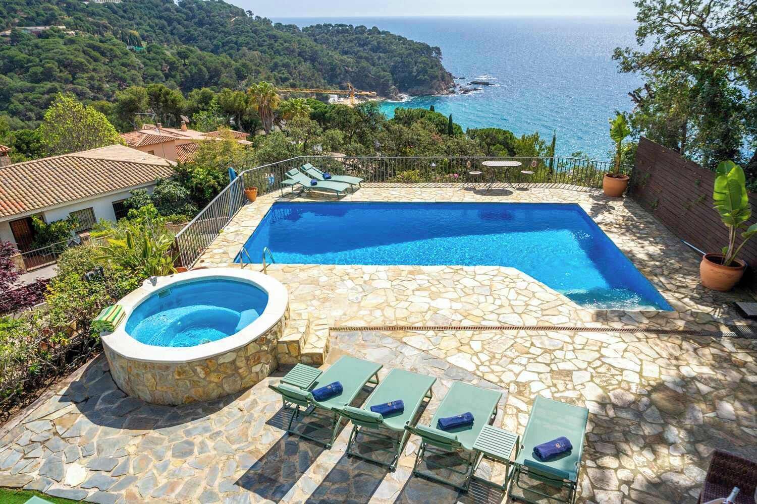 Piscina con vasca idromassaggio, circondata da piastrelle in pietra e lettini, con vista sul paesaggio costiero.
