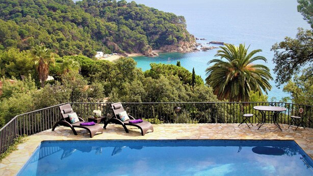 Zona de piscina con sillas de descanso y una mesa, con vistas a un paisaje costero con árboles y al mar.