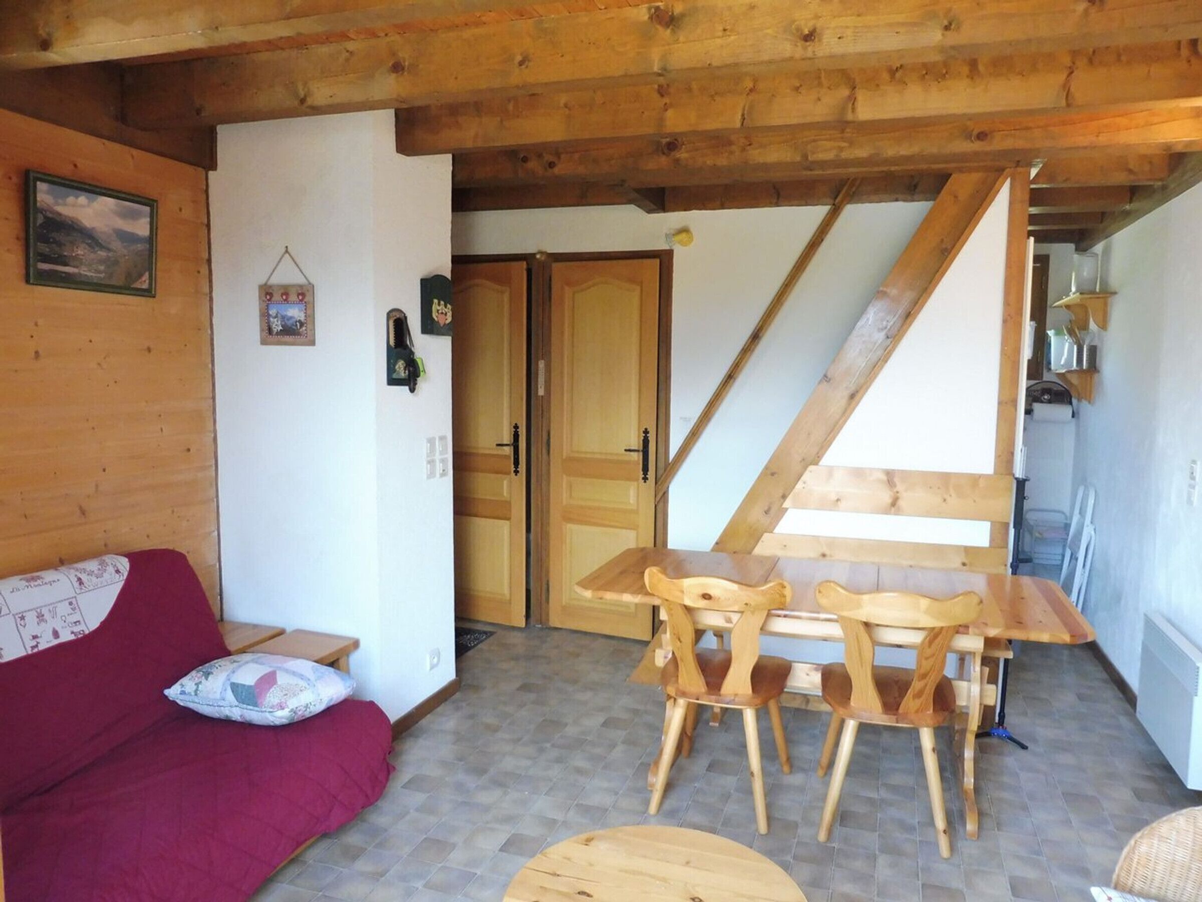 A cozy room with a wooden ceiling, a wooden dining table with chairs, a red sofa with a patterned cushion, and a door leading to another room.