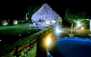 Estrutura com telhado de palha, piscina e cerca de madeira em primeiro plano.