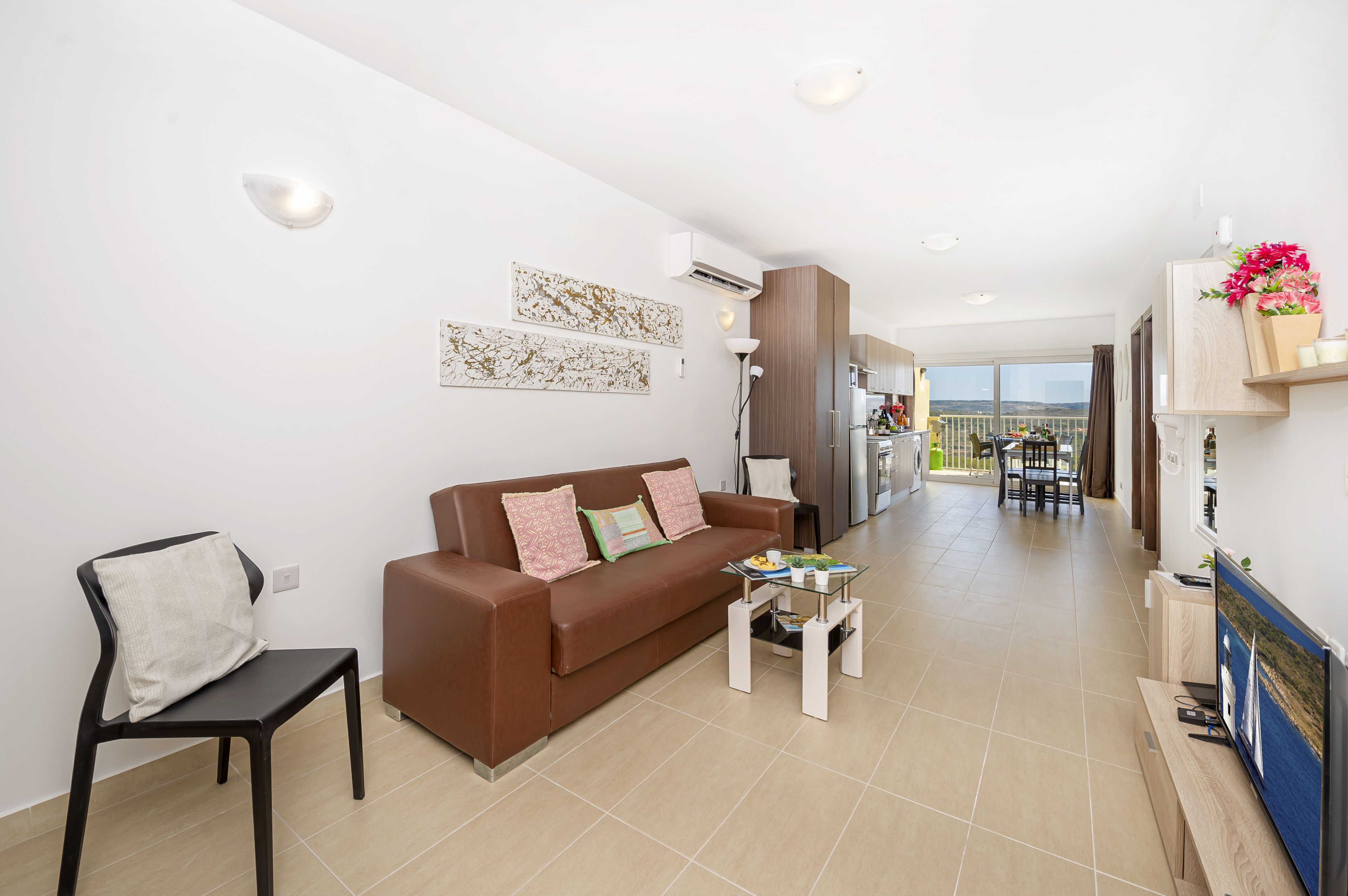 A modern living room with a brown sofa, a small table, and a chair. There is a kitchen in the background with a balcony.