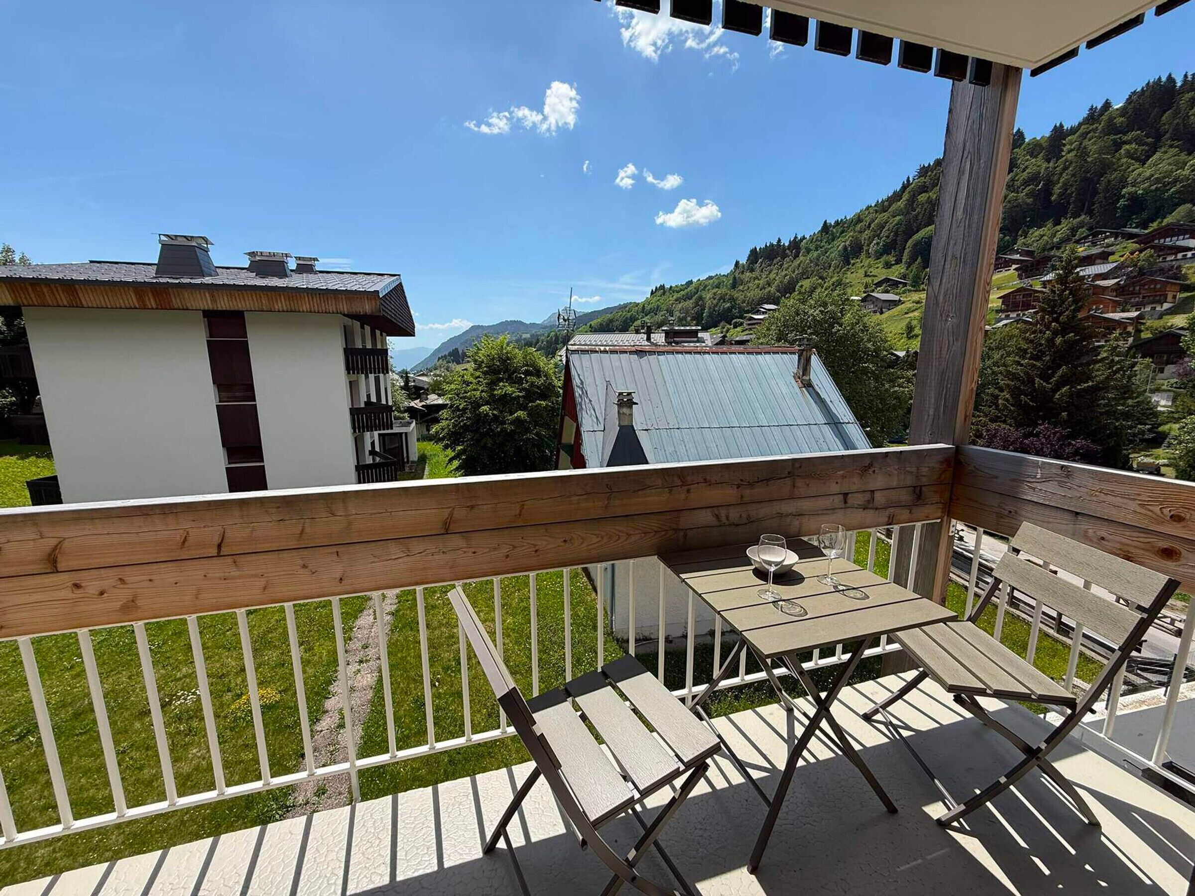 Un balcón de madera con mesa y sillas, con vistas a un paisaje montañoso y casas.
