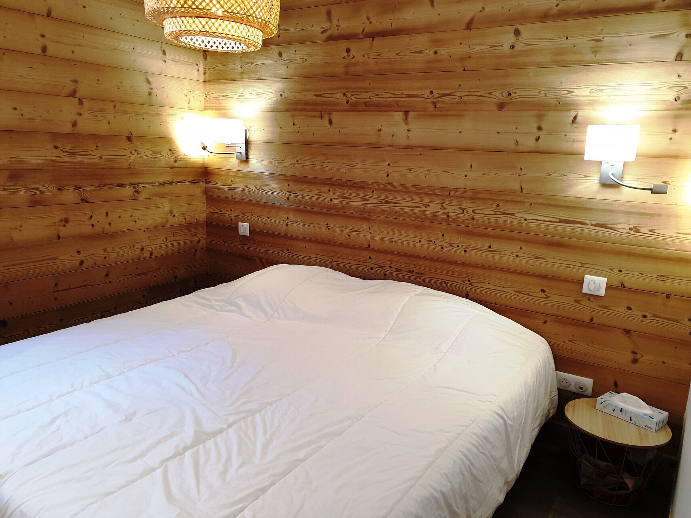 Una habitación con paneles de madera, una cama, dos luces fijadas a la pared y una pequeña mesa redonda con una caja de pañuelos.