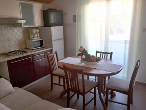 A compact kitchen with a gas stove, microwave, and a small refrigerator. There is a dining table with four chairs and a bowl of fruit. - Guesthouse Raffaello (Rab)