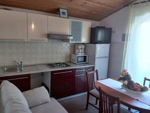 A compact kitchen with a microwave, stove, and refrigerator. There is a dining table with chairs and a small fruit bowl on the table. - Guesthouse Raffaello (Rab)