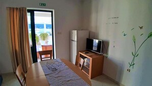 A hotel room with a dining table, chairs, a TV on a wooden cabinet, and a view of the sea through a door. - Guesthouse Raffaello (Rab)