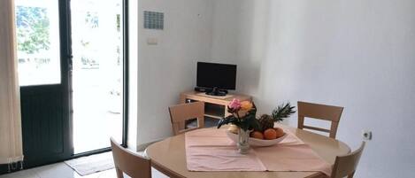 Un comedor con una mesa redonda, cuatro sillas, un televisor pequeño y un frutero con flores sobre la mesa.