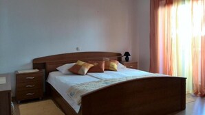 A bedroom with a wooden bed, a nightstand, a lamp, and a window with curtains. - Guesthouse Raffaello (Rab)
