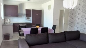 Una sala moderna con un sofá gris, una mesa de comedor con sillas, una zona de cocina con armarios y electrodomésticos, y un papel tapiz estampado en la pared.