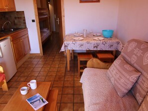 A compact living space with a kitchenette, a dining table set for four, a sofa, and a small table with two mugs and a magazine.