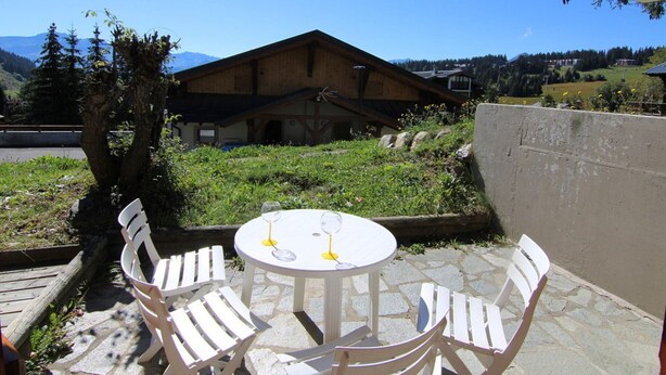 Un patio con sillas blancas y una mesa, un edificio de madera al fondo y un paisaje montañoso.