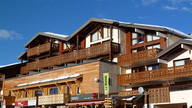 Un immeuble à plusieurs étages doté de balcons en bois, d’un panneau indiquant le site où l’on peut faire du ski, et d’un restaurant appelé « La Bocca ».