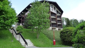 A multi-story building with a sloped roof, surrounded by lush greenery and a pathway leading to the entrance. - Cozy Studio with Balcony, Near Ski Lift, Pool & Tennis, Megève - Sleeps 4 (Megève)
