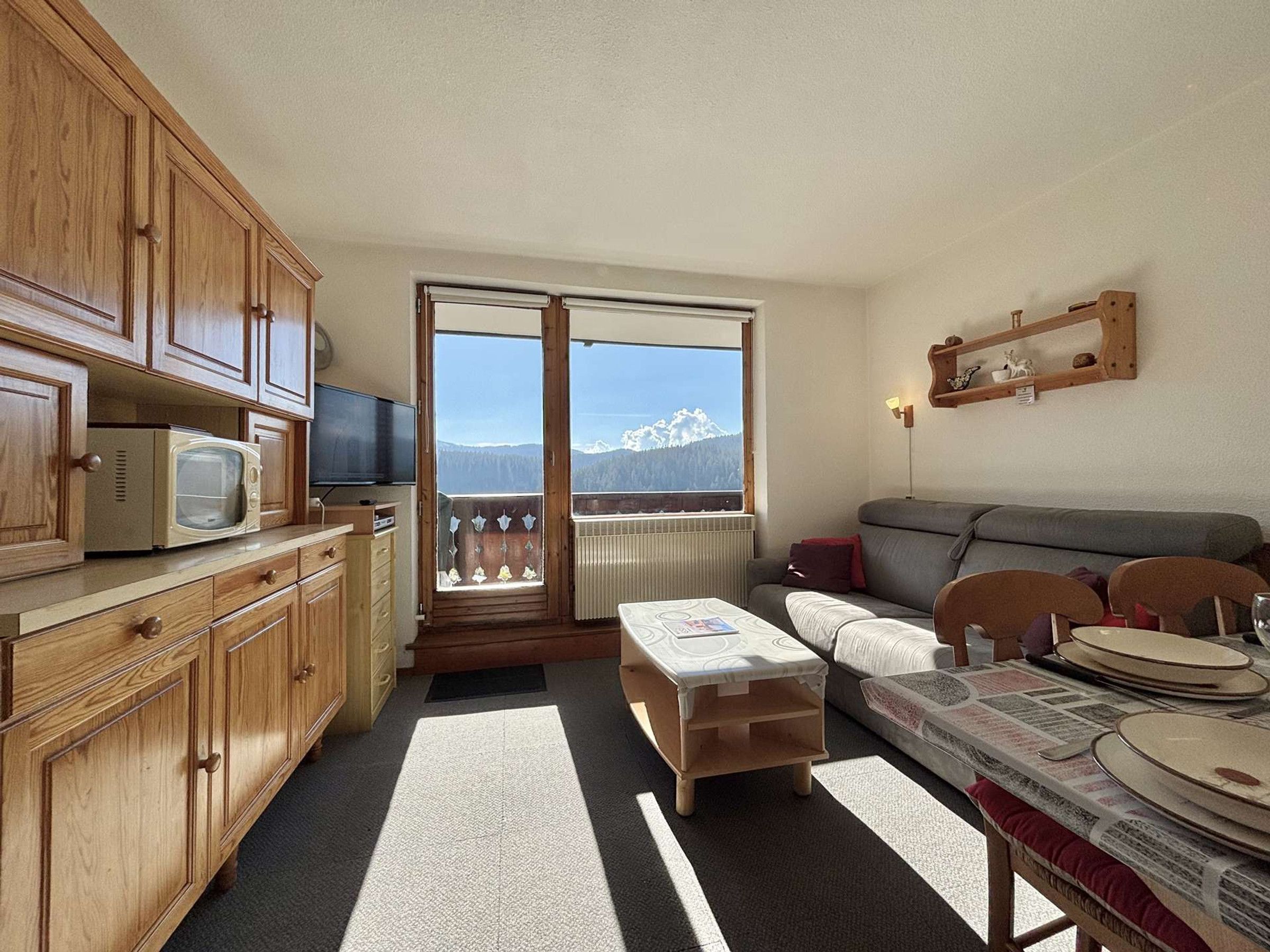 Un espacio habitable compacto con kitchenette, un sofá, una mesa de comedor con platos y vistas a montañas a través de un ventanal grande.