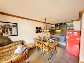 A cozy living room with a sofa and a dining table set for four. A kitchen with a red refrigerator and modern appliances is visible in the background.