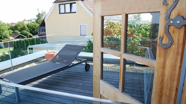 A sun lounger on a wooden deck with a view of a house and greenery. - Alte Schmiede (Bad Griesbach im Rottal)