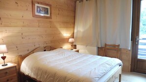 A wooden room with a bed, two bedside tables with lamps, a chair, and a window with curtains. - Cozy 2-Bedroom Apartment with Cabins in Crest-Voland - Near Slopes, Sleeps 6 (Crest-Voland)