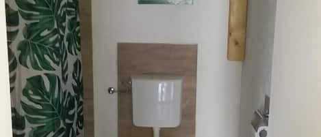 Casa de banho pequena com sanita, cortina de chuveiro com padrão de folha, prateleira de madeira com uma planta e suporte de papel higiénico fixado na parede.