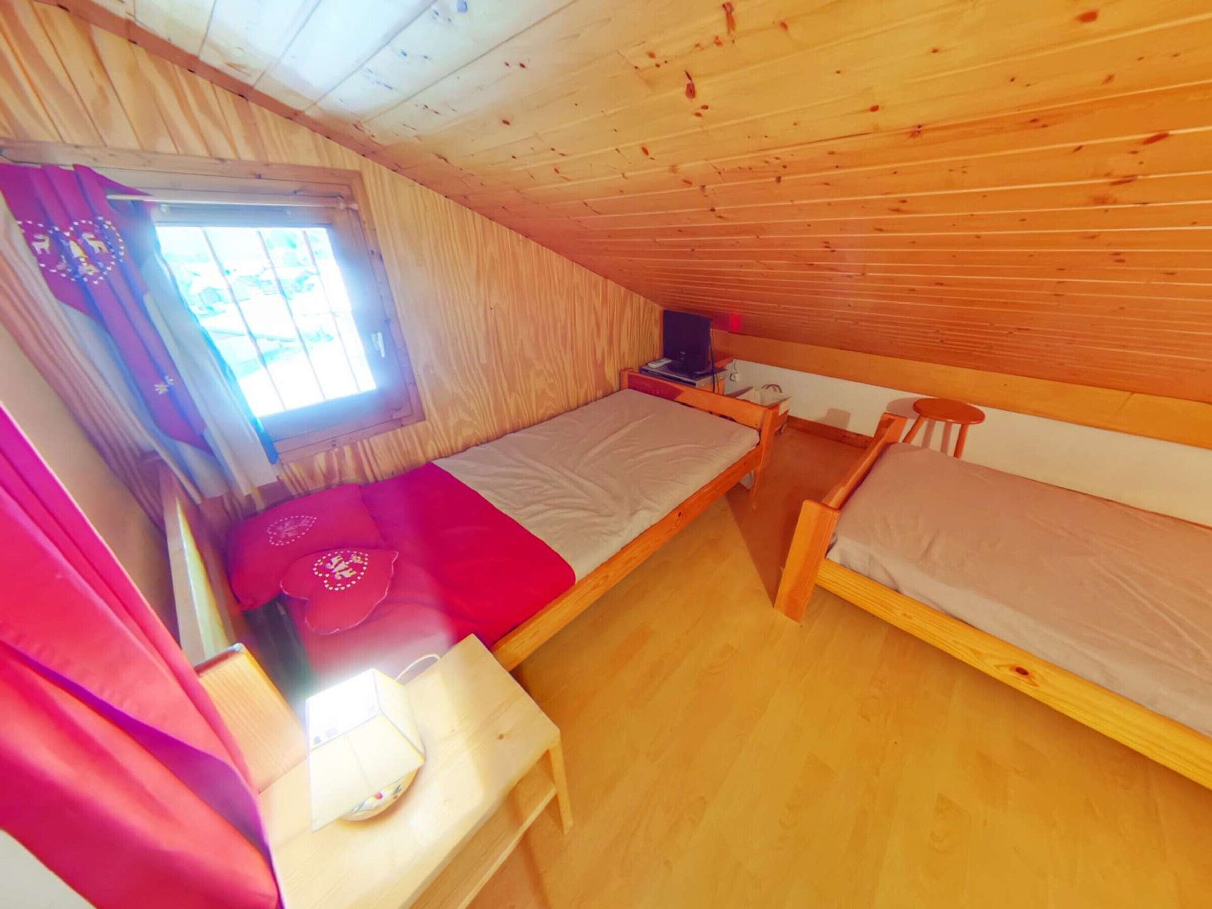 A small, cozy room with wooden walls and a sloped ceiling. There are two single beds, a small table, and a window with curtains.