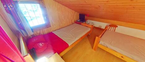 A small, cozy room with wooden walls and a sloped ceiling. There are two single beds, a small table, and a window with curtains.
