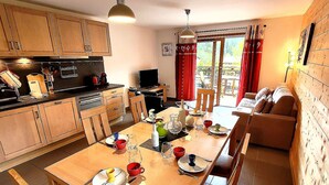 A modern kitchen with wooden cabinets, a dining table set for four, and a large window with red curtains. - Bright 2BR Apartment with Terrace and Parking, Sleeps 4, Les Gets (Les Gets)