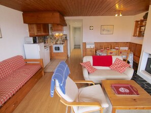 Una sala de estar acogedora con un sofá, una silla y una mesa de centro. Se observa una zona de cocina con armarios de madera y un microondas. Al fondo, se distingue una chimenea.
