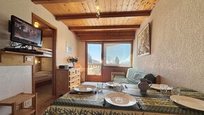 Una habitación acogedora con techo de madera, televisor montado en la pared, cama, mesa de comedor con platos y vasos, y ventana con persianas.