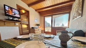 Un comedor con una mesa lista para dos personas, un televisor, un jarrón y una ventana con vista a árboles.
