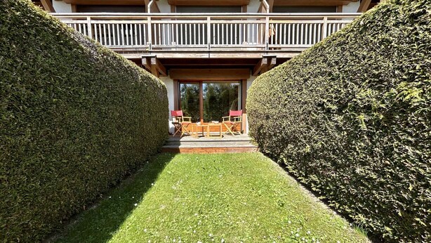 Un sentier bordé de haies soigneusement taillées mène à une terrasse en bois aménagée avec du mobilier d’extérieur.