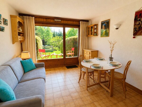 A room with a sofa, a dining table with chairs, a cabinet, and a view of the garden through a sliding door. - Cozy Studio with Alcove, Ski-In Access, Near Lifts & Golf, Sleeps 4 - Megève (Megève)