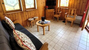 A cozy room with wooden walls, a sofa, a TV, a small table, and two windows with curtains.