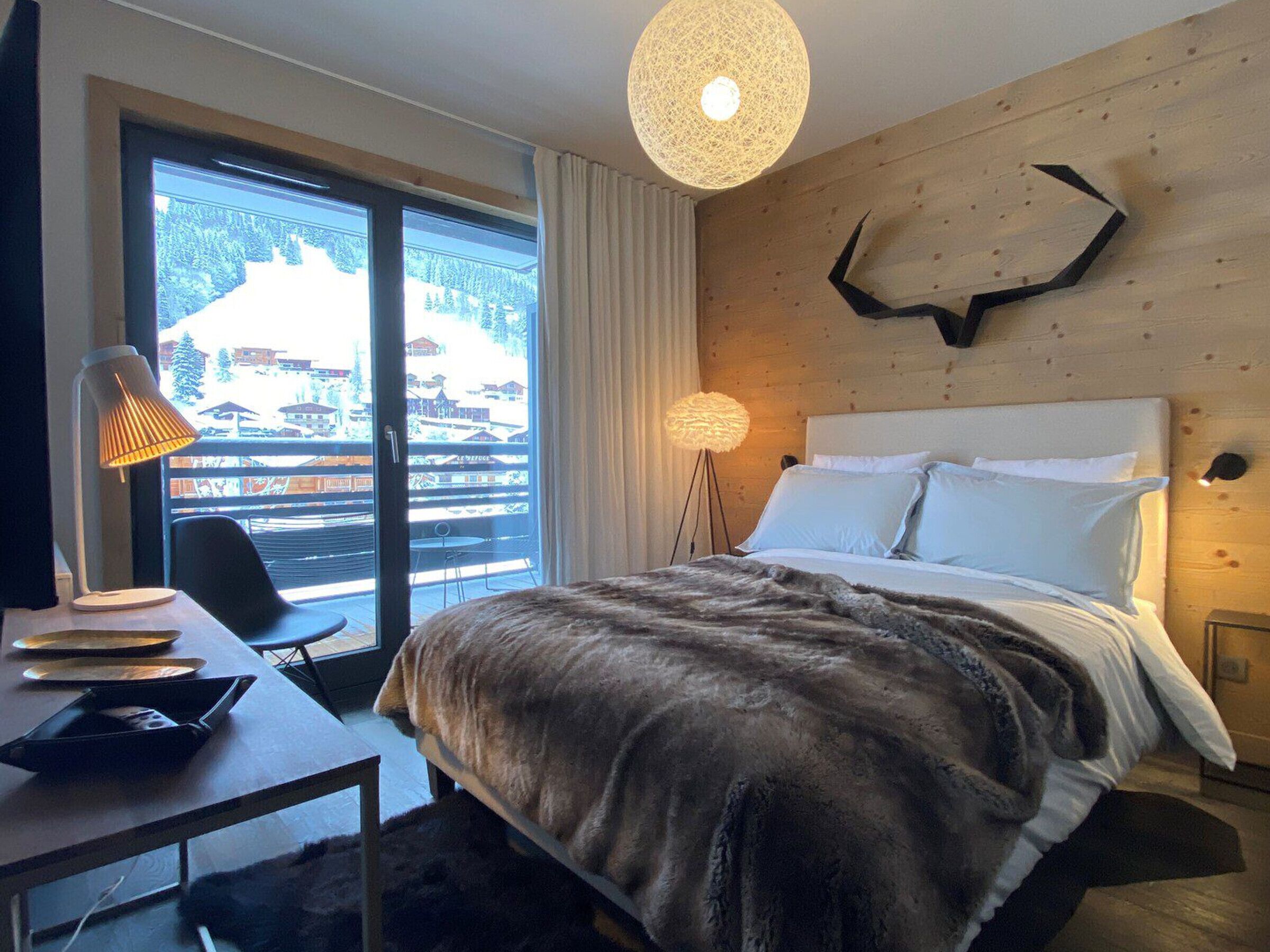 Una camera d'albergo con un letto, una scrivania con lampada, una sedia, un'ampia finestra vista edifici e un'originale decorazione murale.