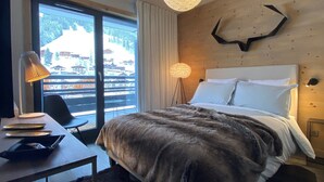 A hotel room with a bed, a desk with a lamp, a chair, a large window with a view of buildings, and a unique wall decoration.