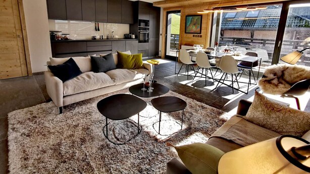 A modern living room with a sofa, coffee table, and chairs. There is a kitchen area with cabinets and a dining table with chairs. The room has large windows and a view of the outside.