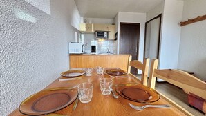 A dining table set with plates, glasses, and cutlery in a room with a kitchenette in the background. - Cozy Studio in Notre-Dame-de-Bellecombe - Ski-in/Ski-out, Sleeps 4, Parking & Balcony (Notre-Dame-de-Bellecombe)