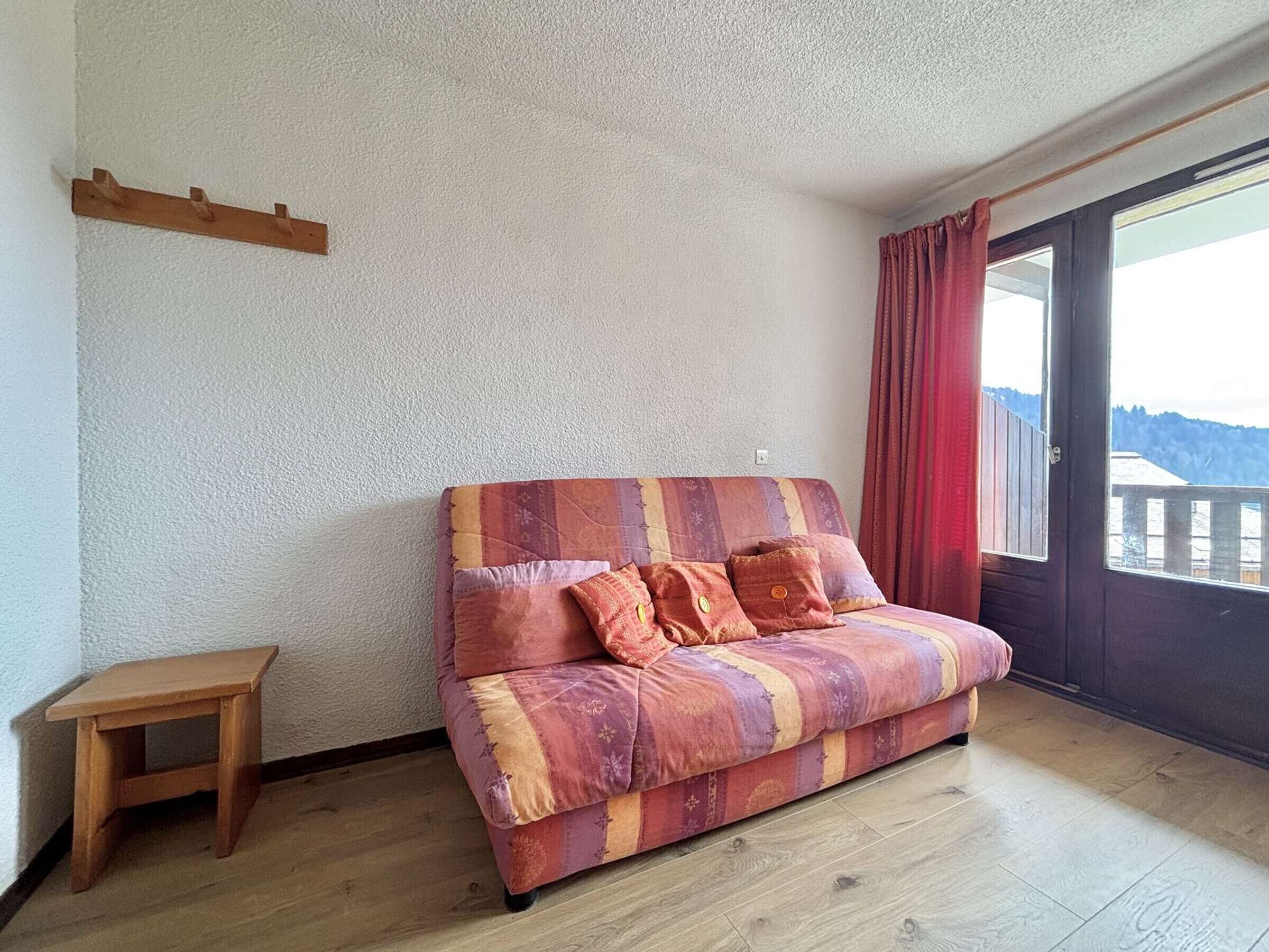 A cozy room with a wooden floor, a small wooden stool, a colorful sofa with cushions, and a large window with a view of a snowy landscape.