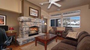 A living room with a stone fireplace, a ceiling fan, a large window with a view of a snowy landscape, a brown sofa, and a wooden coffee table. - Private Lakefront Luxury in the heart of Ski Country, Mountain Views (Silverthorne)